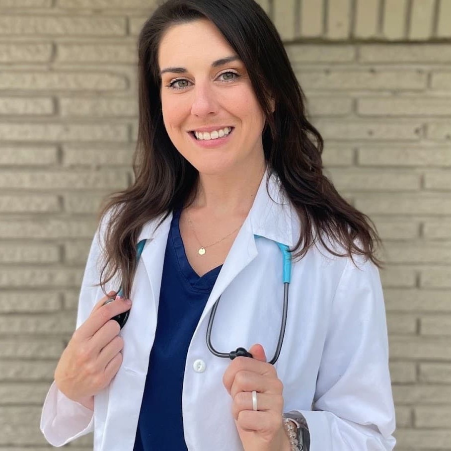 A person wearing a white coat and stethoscope stands in front of a brick wall, smiling.