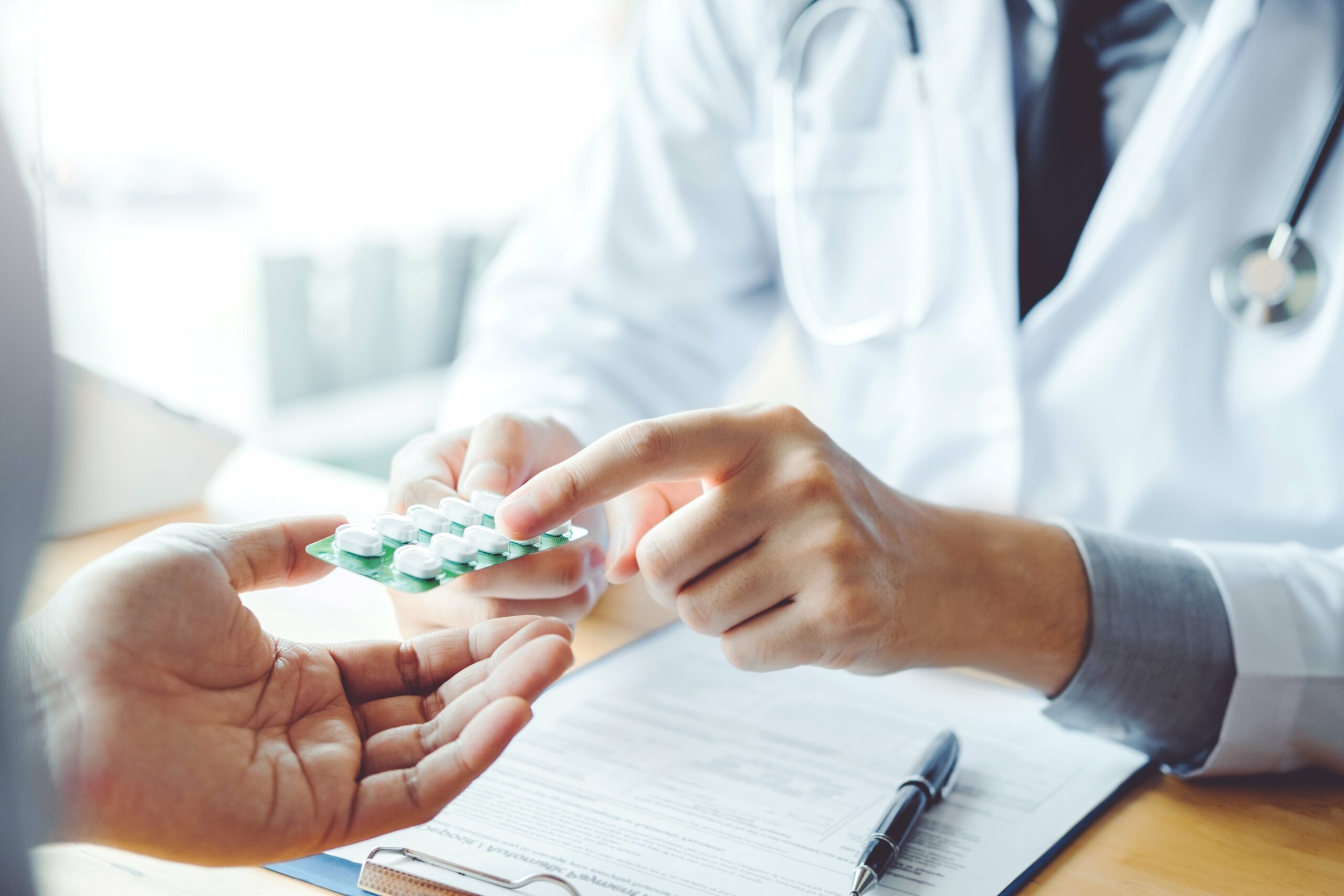 A doctor hands medication to a patient across a desk, reminiscent of a pharmacy setting, with a clipboard and pen visible.