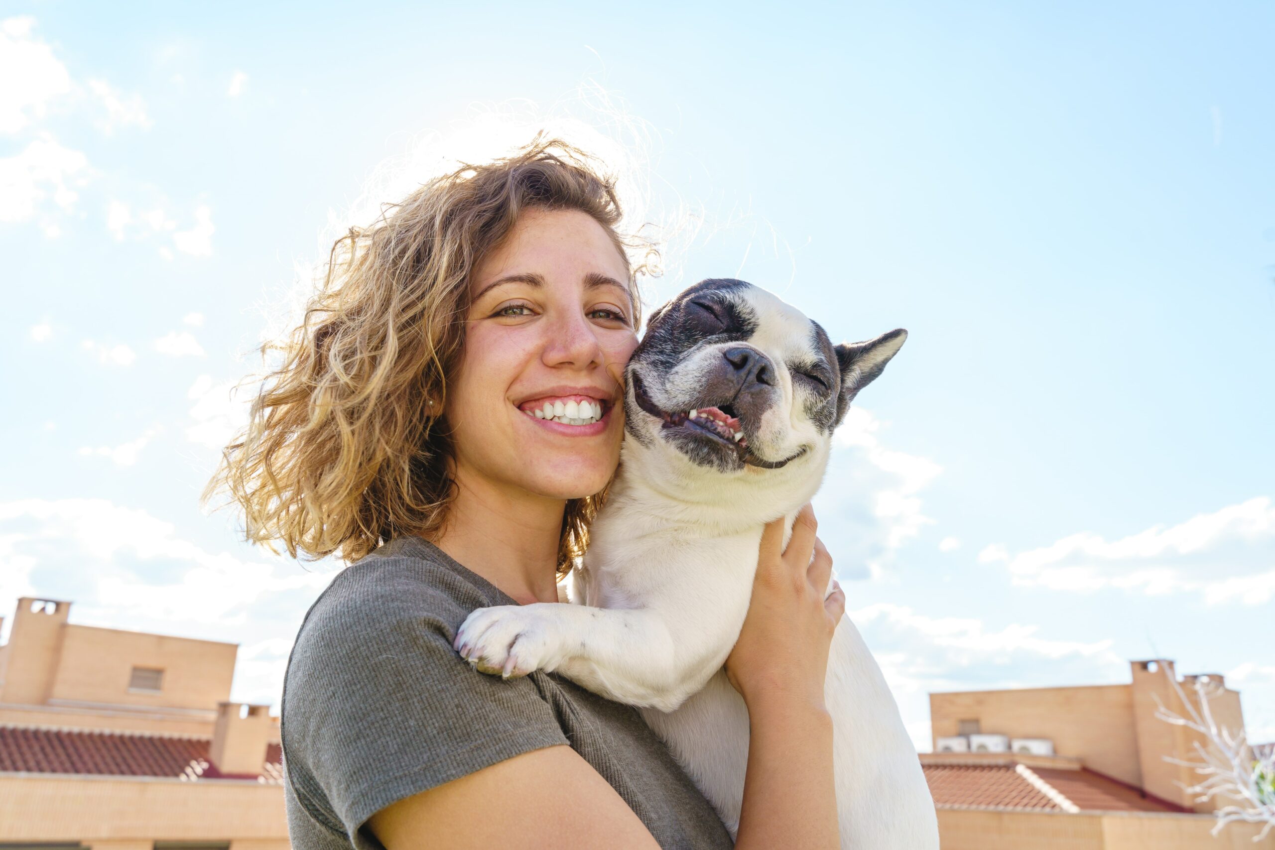 A person with a radiant smile cradles a happy French Bulldog under the bright sun, showcasing the perfect harmony of outdoor joy and good dentistry.