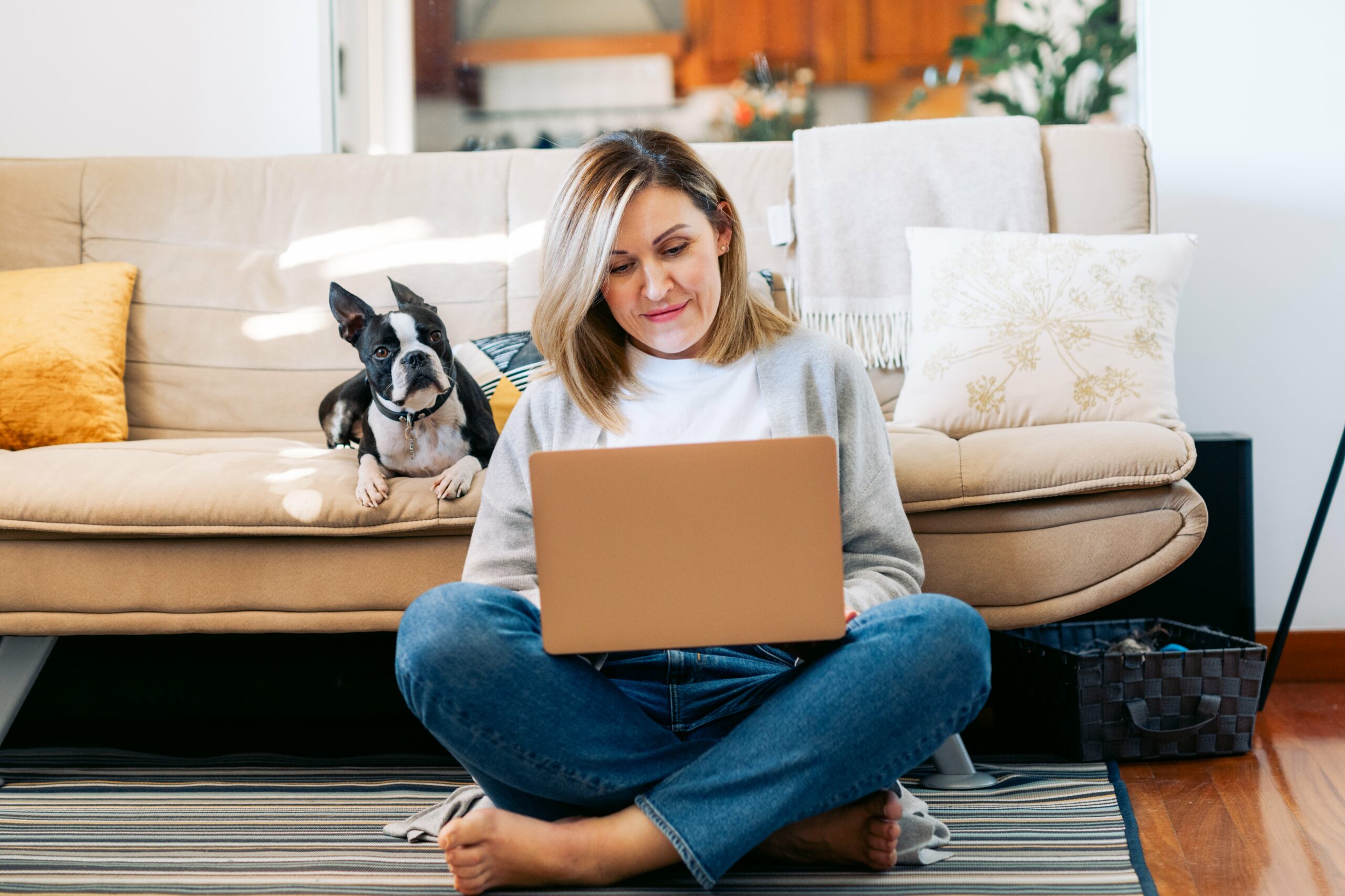 A woman sits cross-legged on the floor using a laptop, with a black and white dog on the sofa behind her in a cozy living room setting.