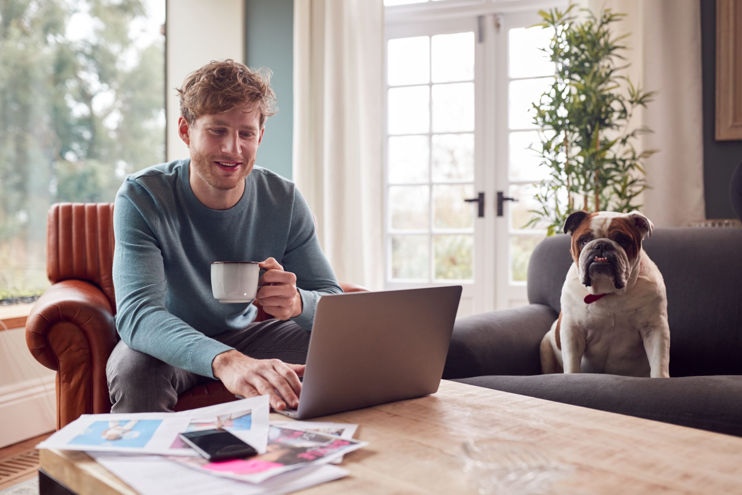 A man relaxes on a couch with his laptop and a coffee cup, while his dog lounges nearby on another couch. Amidst the cozy scene, papers and a phone are strewn across the wooden table, evoking an afternoon spent working on pharmacy research. A window and plants complete the serene backdrop.