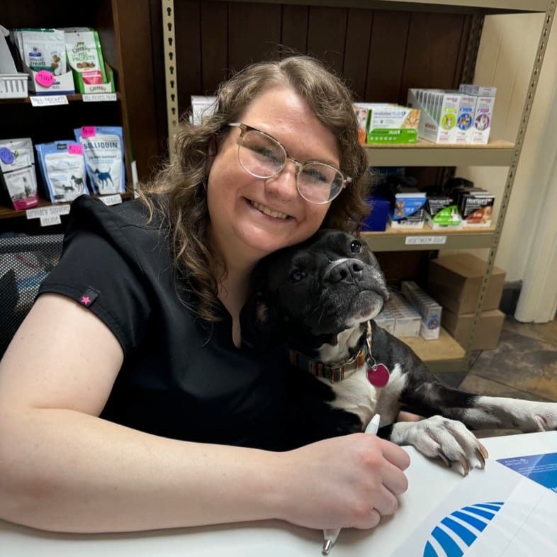 A woman with glasses is seated at a desk, smiling and hugging a black and white dog. Shelves with various products are visible in the background.